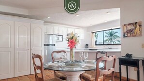 A dining area with a glass table surrounded by wicker chairs, white cabinets, and a kitchen in the background. - Amazing 2 BR Condo Overlooking the Pool - Romantic Zone (Puerto Vallarta)
