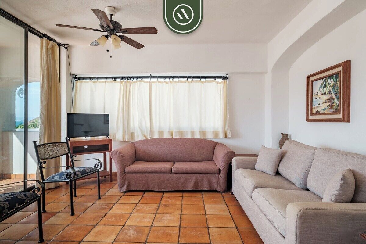 Ein Wohnzimmer mit einer Couch, einem Fernseher, einem Deckenventilator und einem Fenster mit Vorhängen.