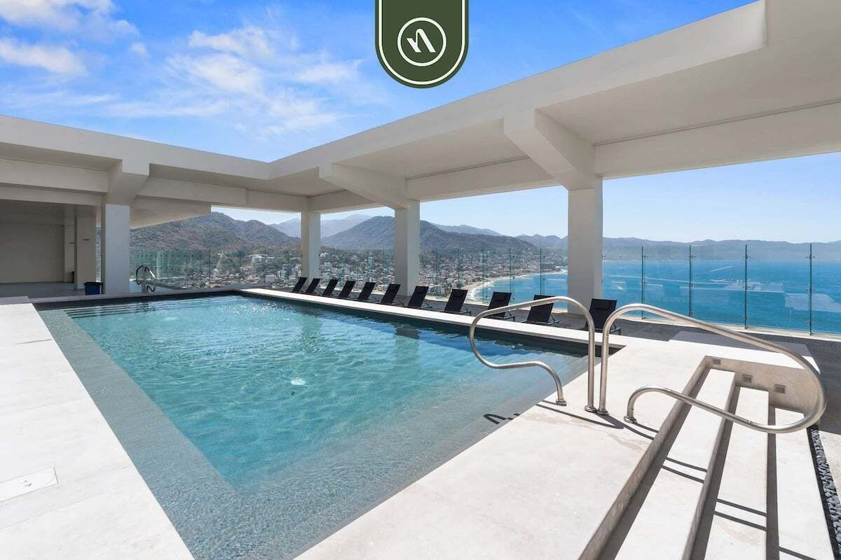 A modern indoor swimming pool with clear blue water, surrounded by white pillars and a view of a coastal city and mountains in the background.