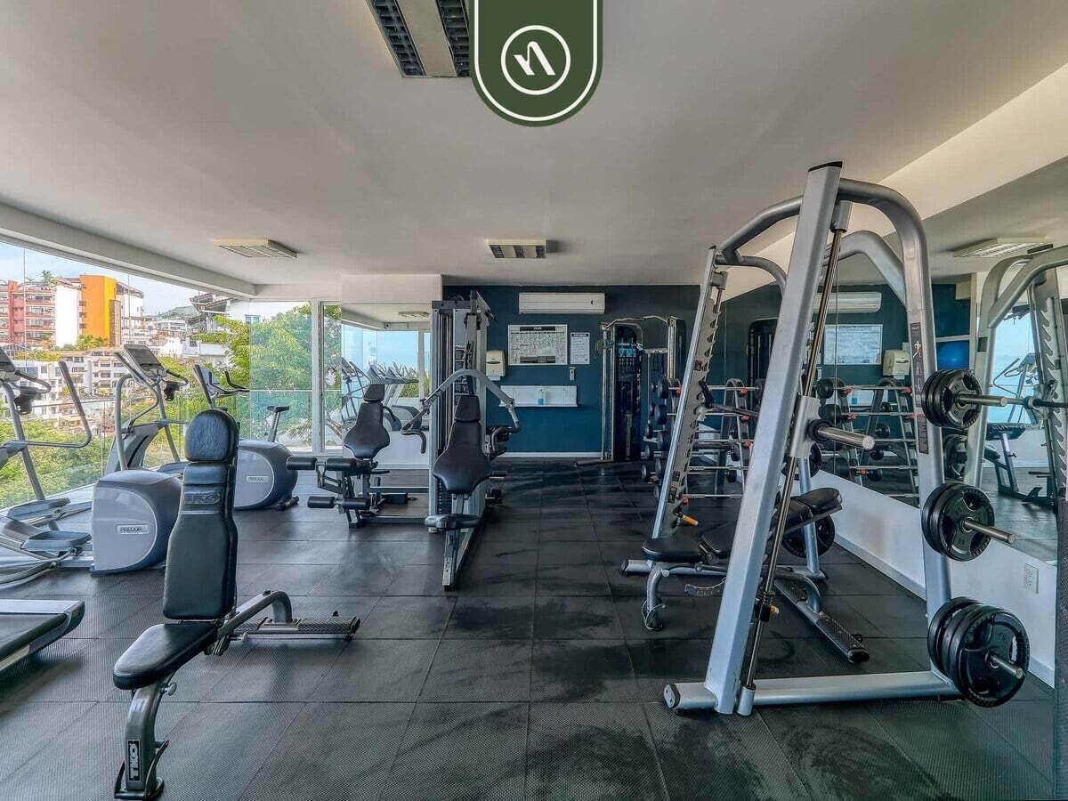 Un gimnasio con equipo de ejercicio, una unidad de aire acondicionado montada en la pared y una ventana con vistas a edificios.