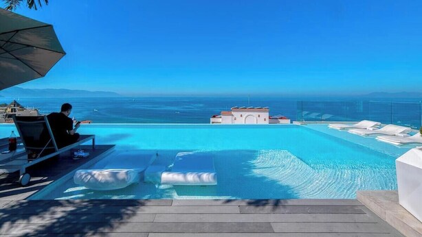 A pool with a clear view of the sea, a lounge area with chairs, and a building in the background.