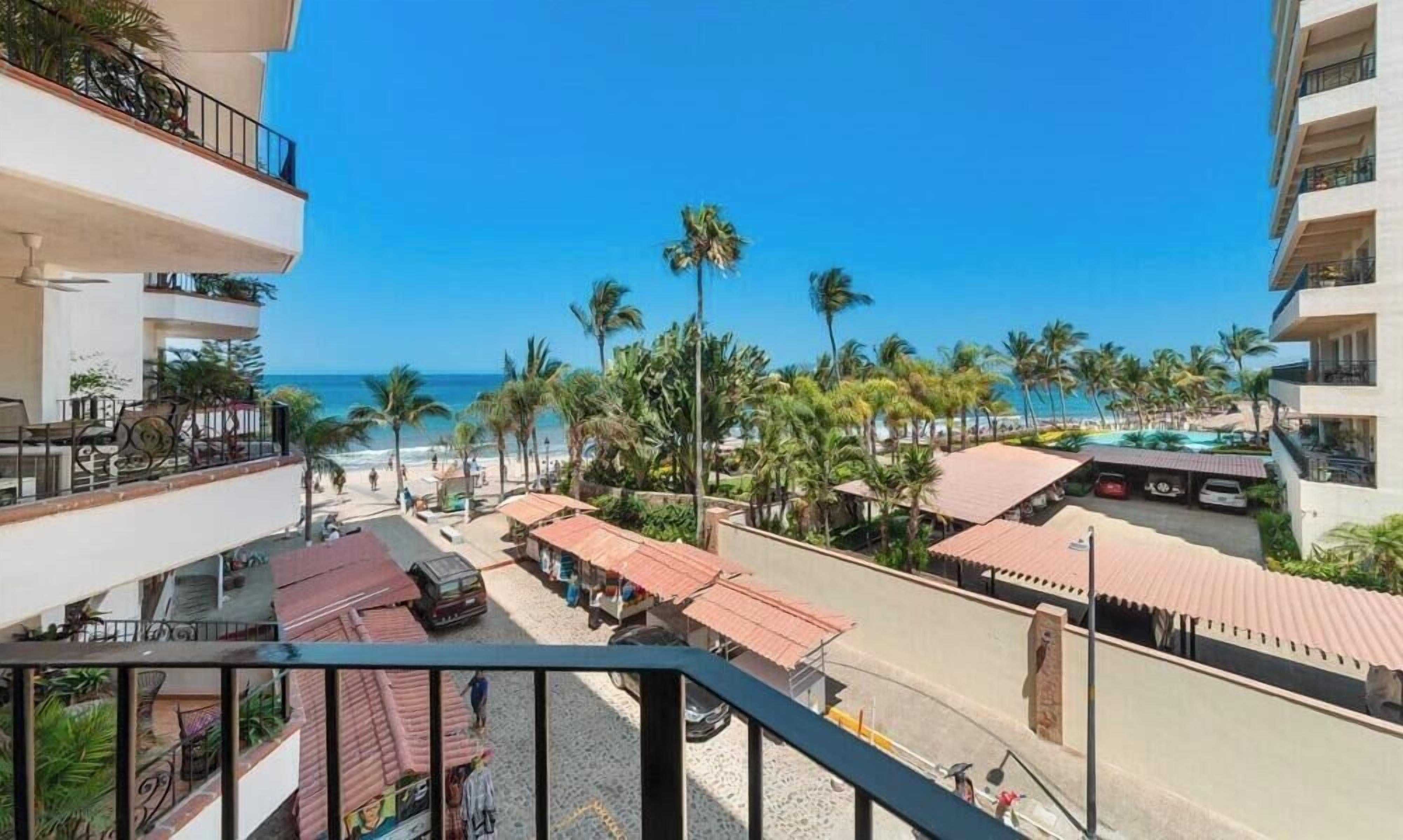 A hotel with balconies overlooking a beach, a pool area, and a street with parked cars.