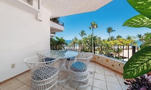 A balcony with a glass table and wicker chairs, overlooking a beach with palm trees and the ocean. - Ocean View 2Br Condo-balcony-beach-pool - 305 (Puerto Vallarta)