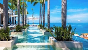 An infinity pool with lounge chairs and palm trees, leading to a beach view. - Luxury 1 BR Condo in Pier 57 - Infinity Pool (Puerto Vallarta)