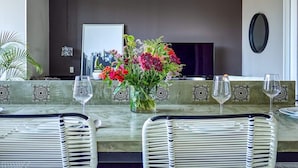 A dining area with a long table, chairs, a vase of flowers, and a television in the background. - Luxury 1 BR Condo in Pier 57 - Infinity Pool (Puerto Vallarta)