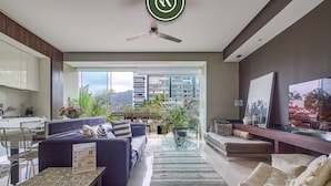 A modern living room with a sofa set, a coffee table, and a TV. There is a balcony with plants and a view of buildings. - Luxury 1 BR Condo in Pier 57 - Infinity Pool (Puerto Vallarta)
