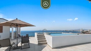A rooftop pool area with a view of the sea, outdoor seating, and a staircase leading to the pool.