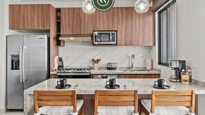 A modern kitchen with wooden cabinets, a stainless steel refrigerator, a microwave, a stove, and a marble countertop. There are three wooden chairs with cushions.
