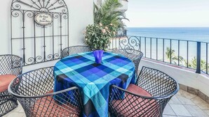 Un balcon avec une nappe à carreaux, des chaises en métal, une grille décorative en métal et une vue sur la mer.