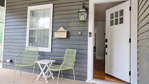 A porch with two green metal chairs and a small table, a white door, and a wall-mounted lantern.