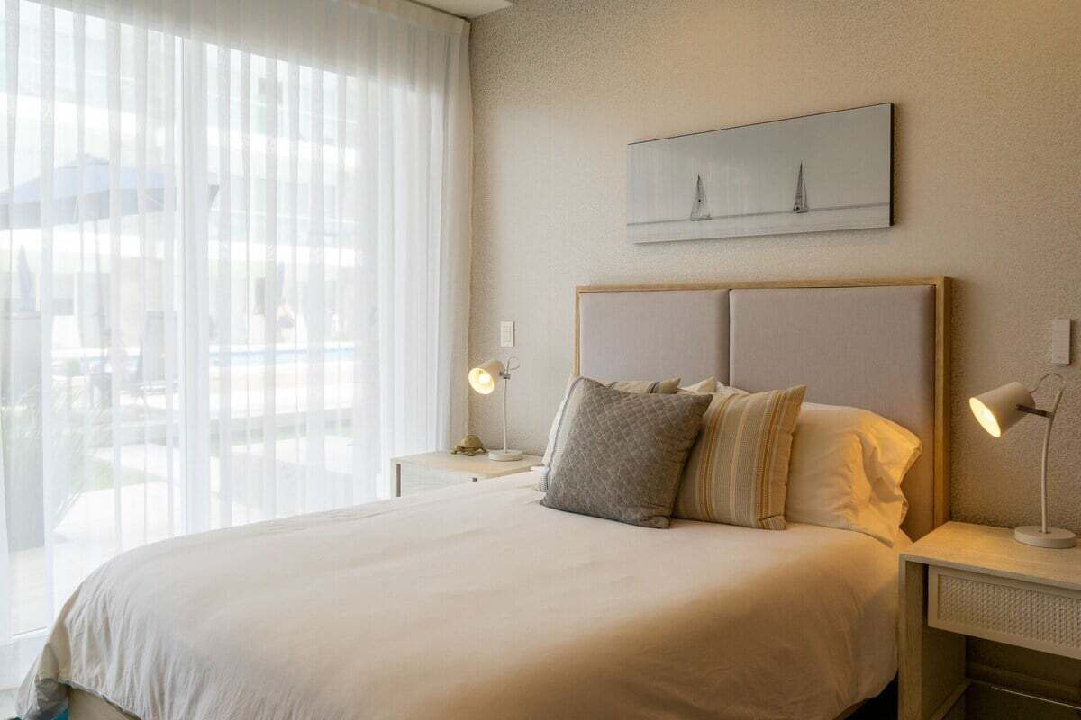 A hotel room with a large bed, two bedside tables with lamps, a window with sheer curtains, and a wall-mounted artwork.