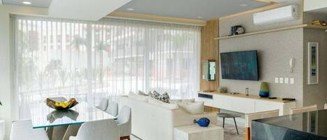 A modern living room with a glass dining table, white sofa, and a flat-screen TV mounted on the wall.