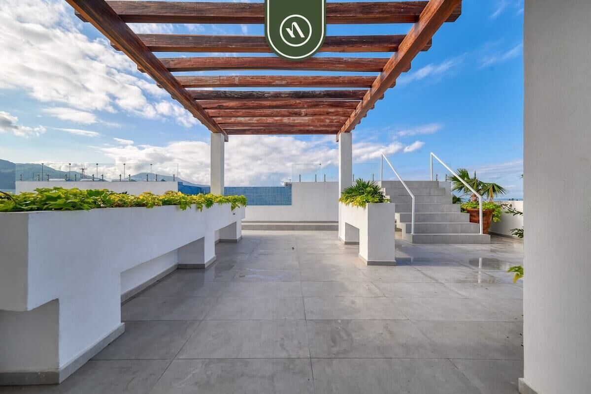 A rooftop terrace with a wooden pergola, white planters with greenery, and a view of the cityscape.