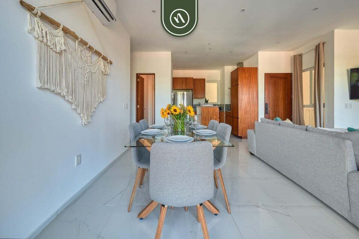 A modern dining area with a glass table, grey upholstered chairs, and a macrame wall hanging.