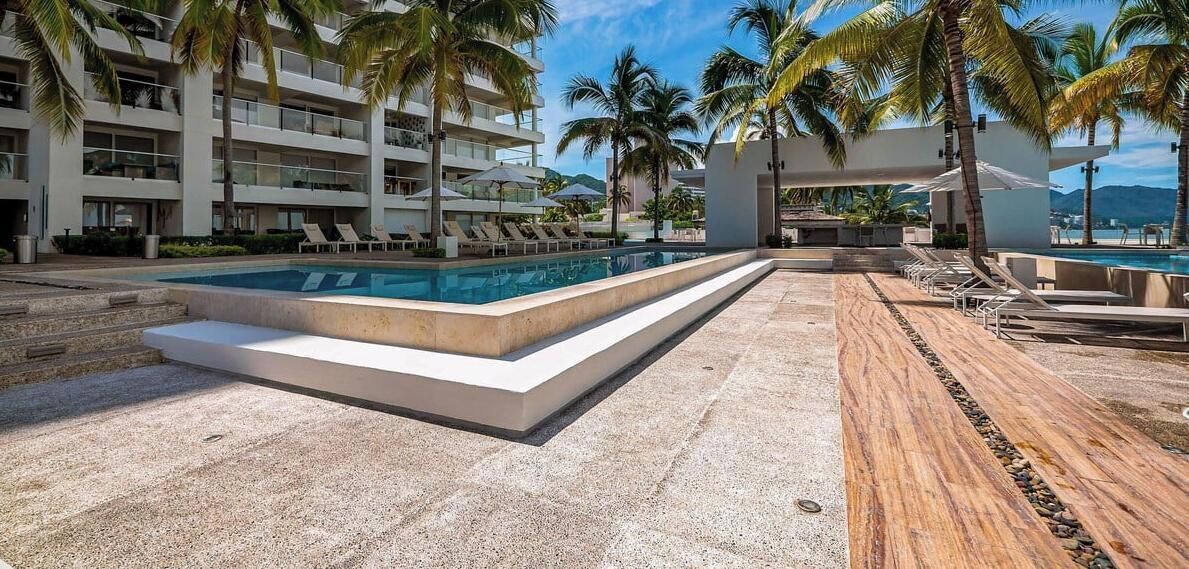 A resort pool area with a rectangular pool, lounge chairs, palm trees, and a multi-story building in the background.