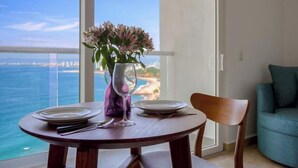 A dining table with plates, a wine glass, and a vase of flowers, overlooking a beach and ocean. - Oceanview Studio - King Bed - Pools - Beachclub (Puerto Vallarta)