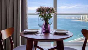 A dining table with two chairs, a vase with flowers, and a view of the ocean and city skyline. - Oceanview Studio - King Bed - Pools - Beachclub (Puerto Vallarta)