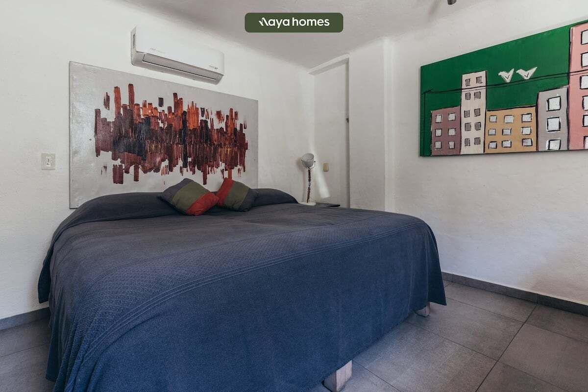 A bedroom with a bed, a wall-mounted air conditioner, a floor lamp, and a cityscape painting.