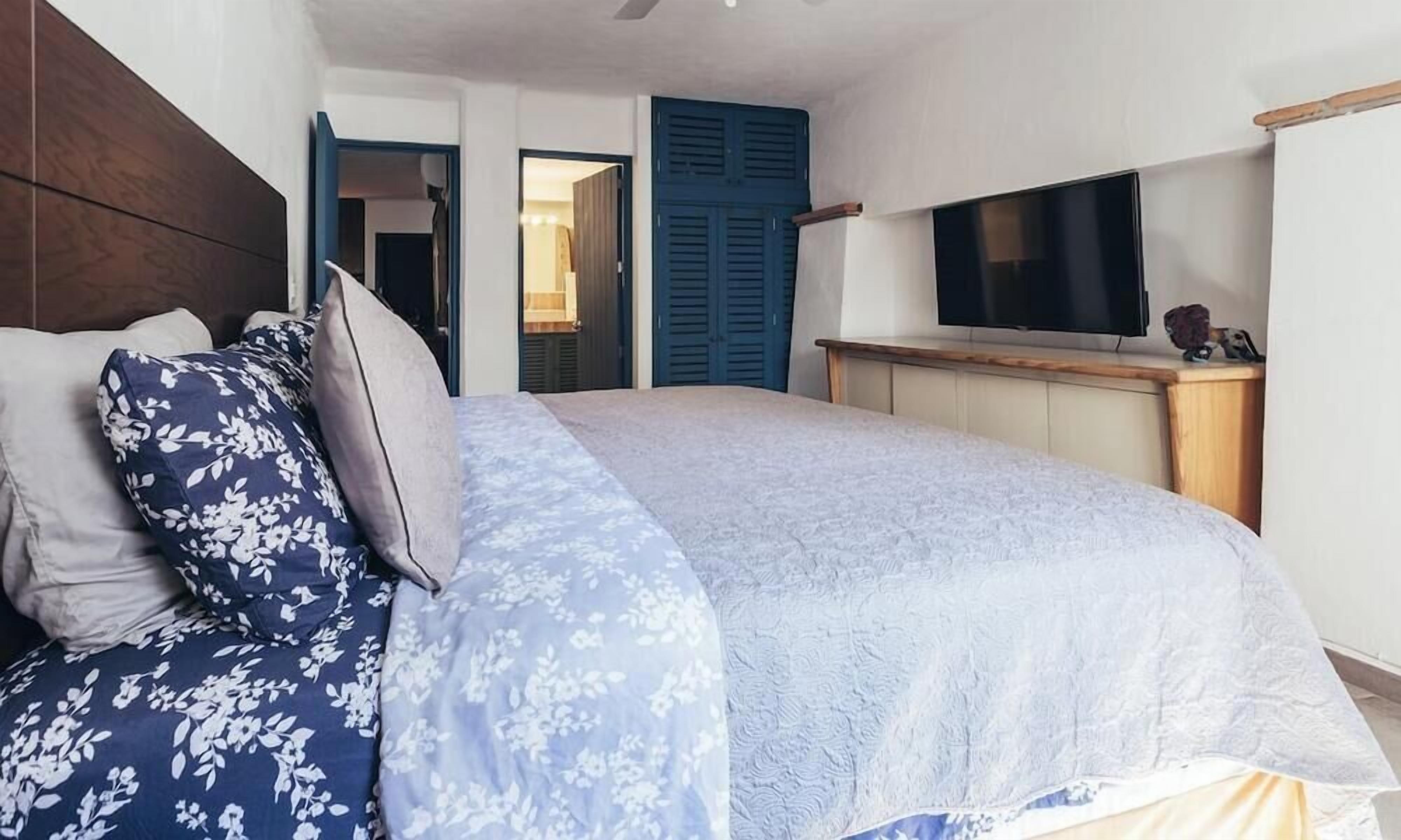 A bedroom with a bed, a TV on a wooden stand, and a door leading to another room.
