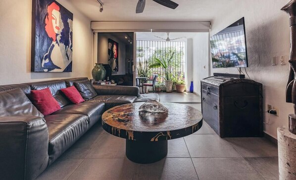 A living room with a leather sofa, a round coffee table, a television, and a large painting on the wall. - Cozy 2BR King Beds - Rooftop Pool - Fast Wifi (Puerto Vallarta)