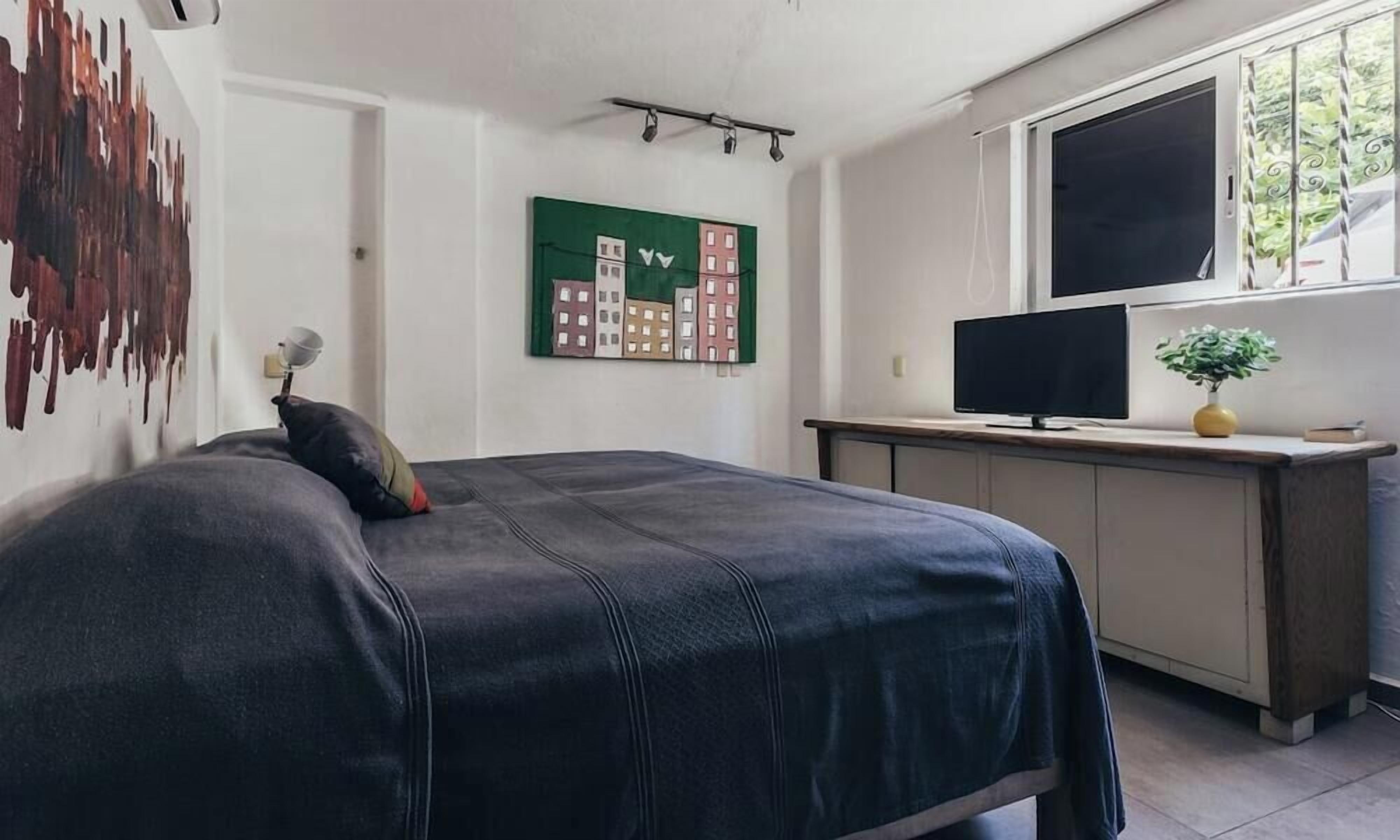A bedroom with a bed, a desk, a television, and a painting on the wall.