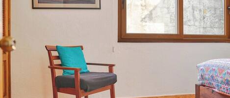 A room with a wooden chair, a bed with a patterned bedspread, a framed picture on the wall, and a window with a view of a building.