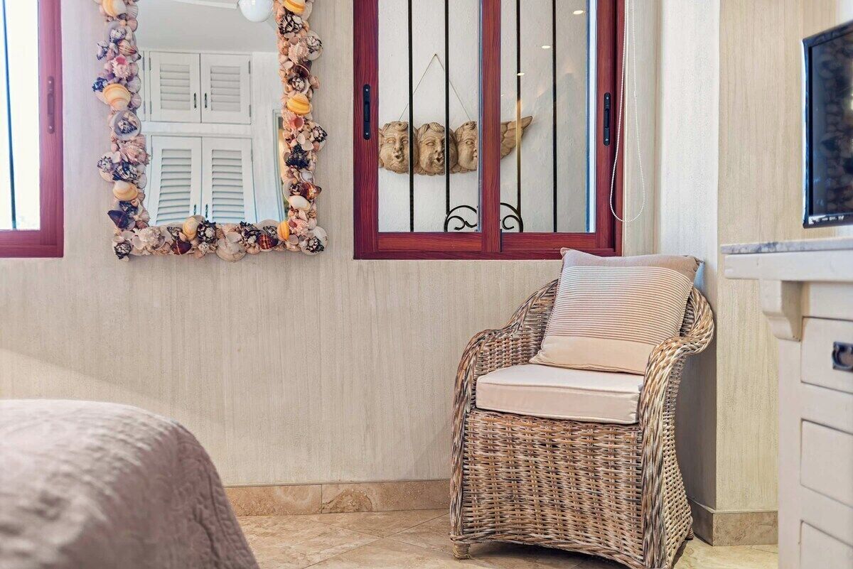 Une chaise en osier avec un coussin, un miroir orné d’une bordure de coquillages et une fenêtre avec une grille en métal.