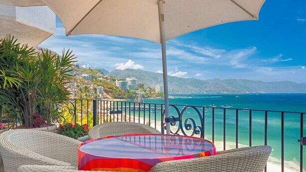 A balcony with wicker furniture and a table, overlooking a coastal view with buildings and mountains. - Ocean View 2Br Condo-balcony-beach-pool - 712 (Puerto Vallarta)