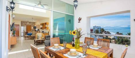 A dining area with a large table set for six, a vase with flowers, and a view of the ocean.