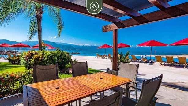 Un área de estar al aire libre con mesas y sillas de madera, sombrillas rojas y vistas a la playa y las montañas.