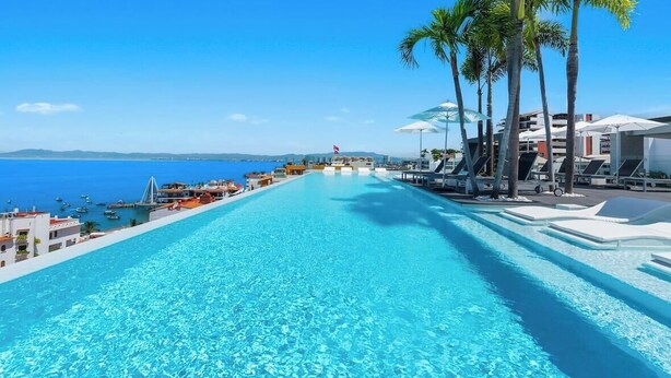 Una piscina con agua cristalina azul, rodeada de sillas de descanso y sombrillas, con vista al mar y edificios al fondo.