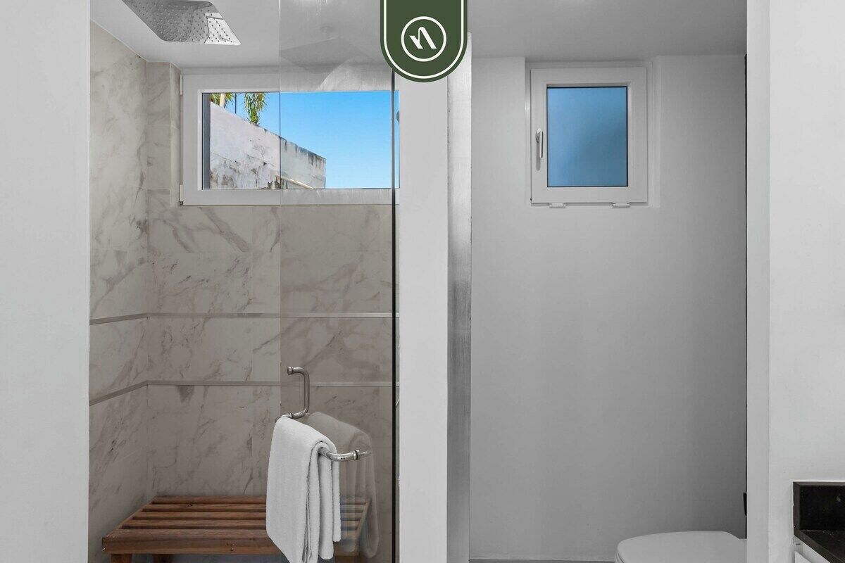 A modern bathroom with a glass shower enclosure, a small window, and a wooden bench.