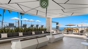 Un área de estar moderna al aire libre con bancos geométricos blancos, sombrillas y vista al mar.