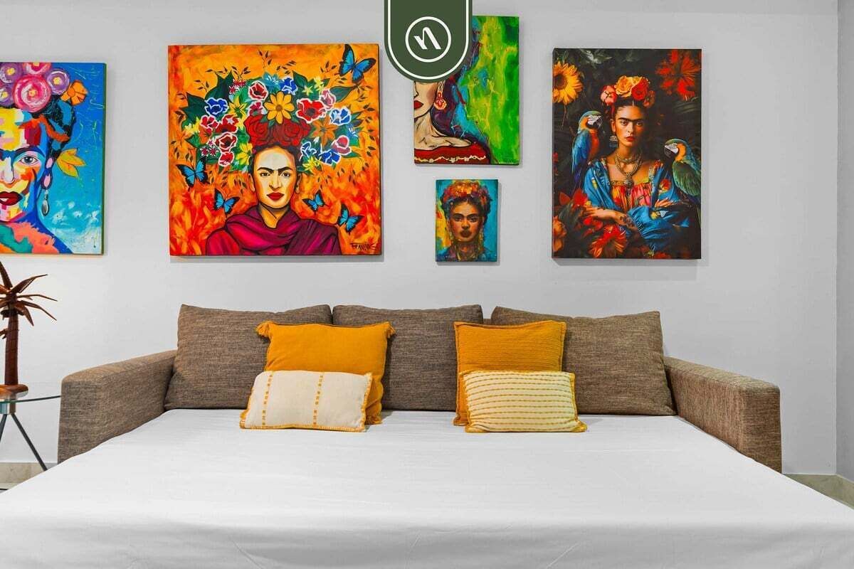 A bed with a white sheet, a brown sofa with two pillows, and four colorful paintings on the wall.