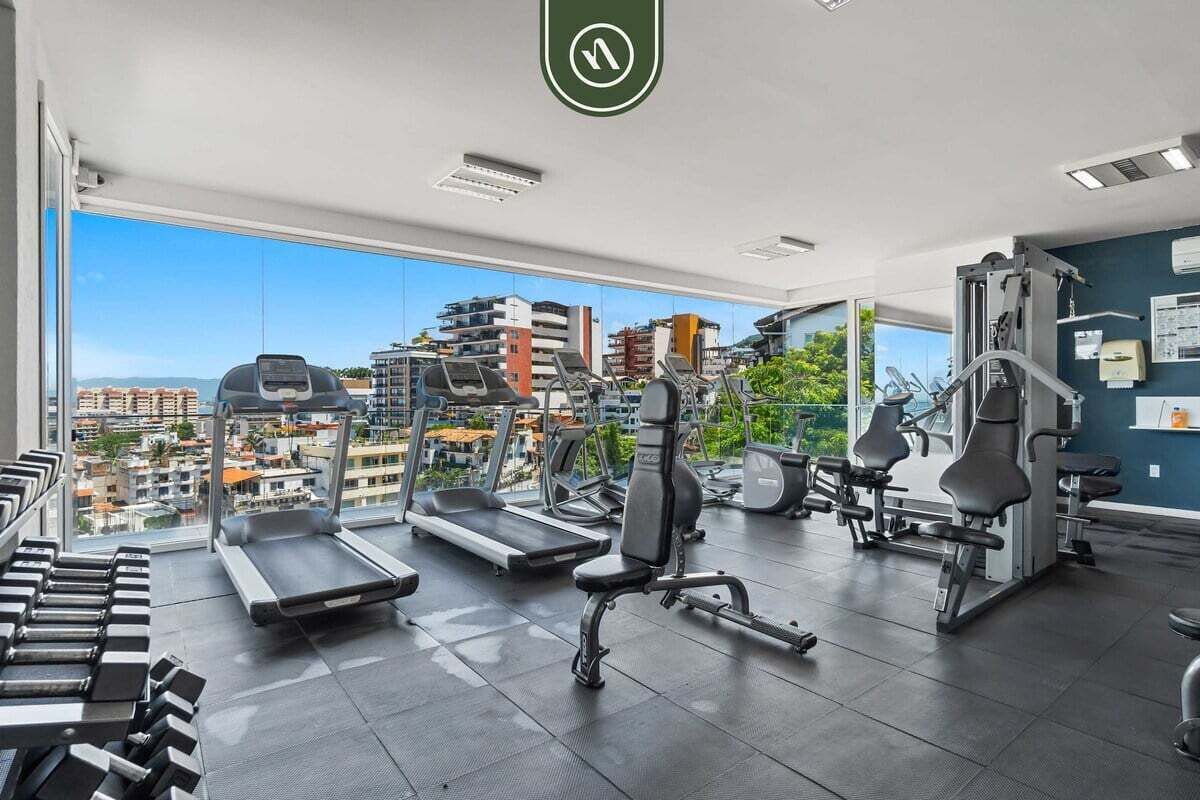 A gym with treadmills, dumbbells, and weight machines, featuring large windows with a city view.