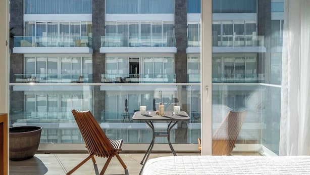 A modern hotel room with a balcony, a dining table with chairs, and a large window offering a view of the building's interior.