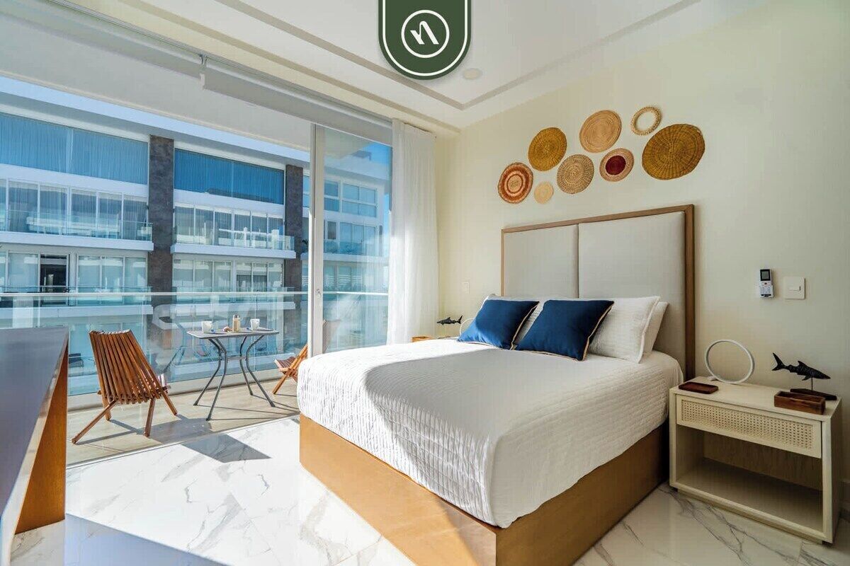 A modern hotel room with a large bed, a balcony with a view of the city, a small table, and a chair.