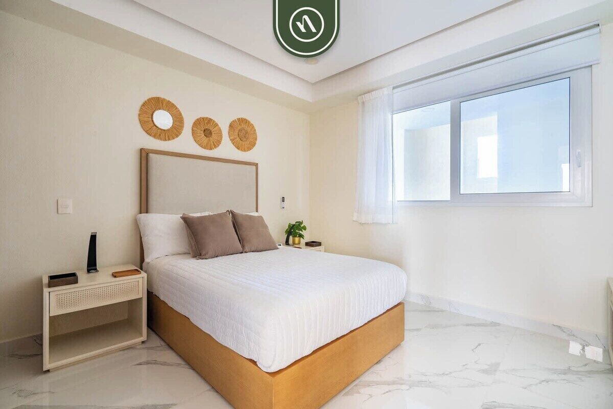 A bedroom with a bed, a nightstand, a wall-mounted mirror, and a window with curtains.