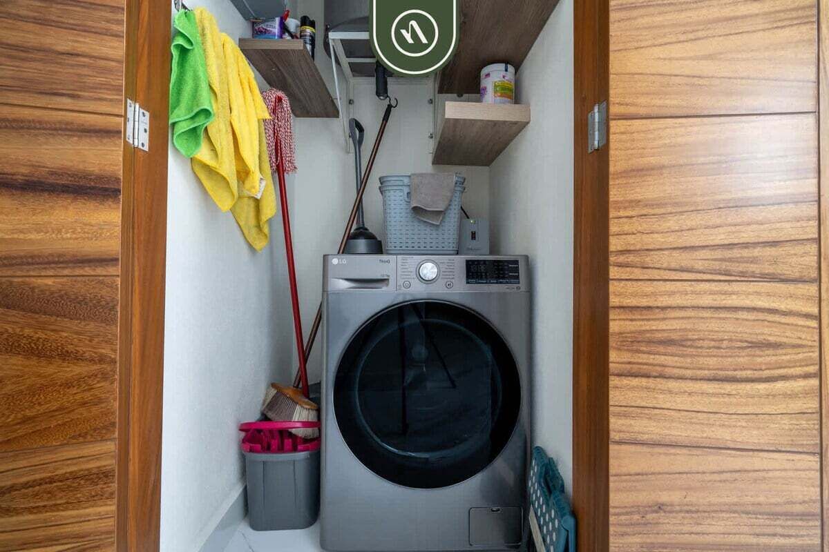 Un espace réduit dédié à la buanderie, comprenant un lave-linge à chargement frontal, un balai et une poubelle.