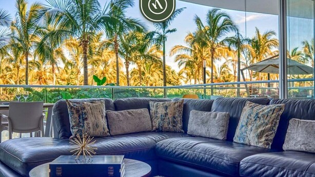 A modern living room with a leather sectional sofa, a round coffee table, and a view of palm trees through large windows.