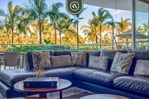 A modern living room with a leather sectional sofa, a coffee table, and a view of palm trees through large windows. - Oceanfront 2BR Condo - Pool - Cevicheria - Beach (Nuevo Nayarit)