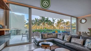 A modern living room with a leather sofa, two round coffee tables, and a large sliding glass door leading to a balcony with palm trees.