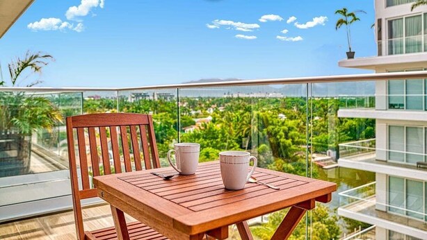 Un balcón con una mesa de madera y dos tazas blancas, con vistas a un paisaje verde exuberante y un paisaje urbano.