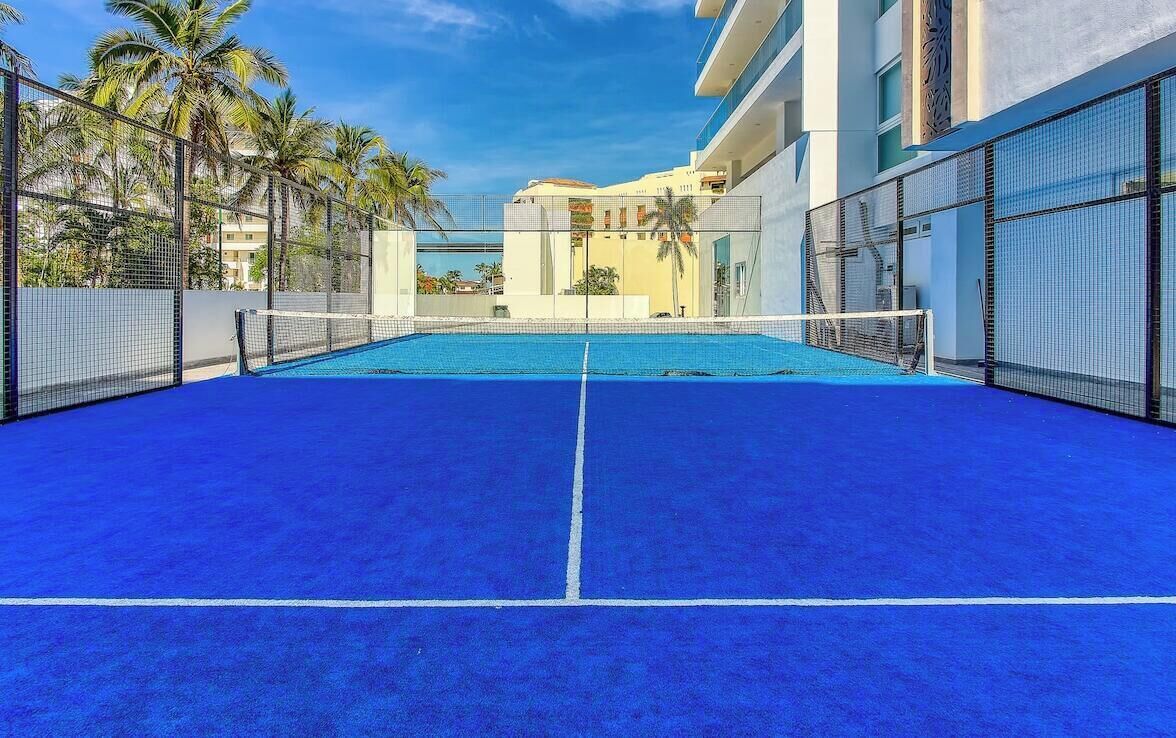 A tennis court with a net, surrounded by a fence, palm trees, and a building in the background.
