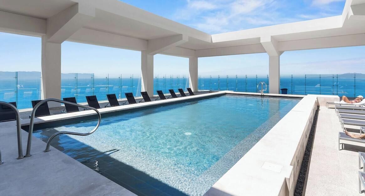 A rooftop infinity pool with clear blue water, white poolside chairs, and a view of the ocean.