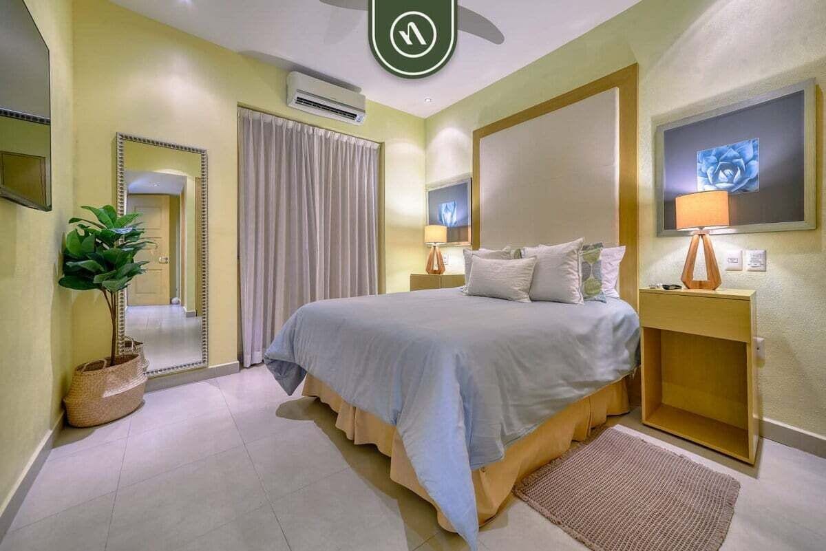 A hotel room with a bed, bedside tables, a mirror, a potted plant, and a painting on the wall.