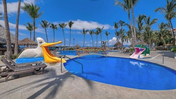 A pool area with lounge chairs, a bridge, and a slide, surrounded by palm trees. - Amazing 2BR Condo in Nuevo Vallarta - Pool - King (Nuevo Nayarit)