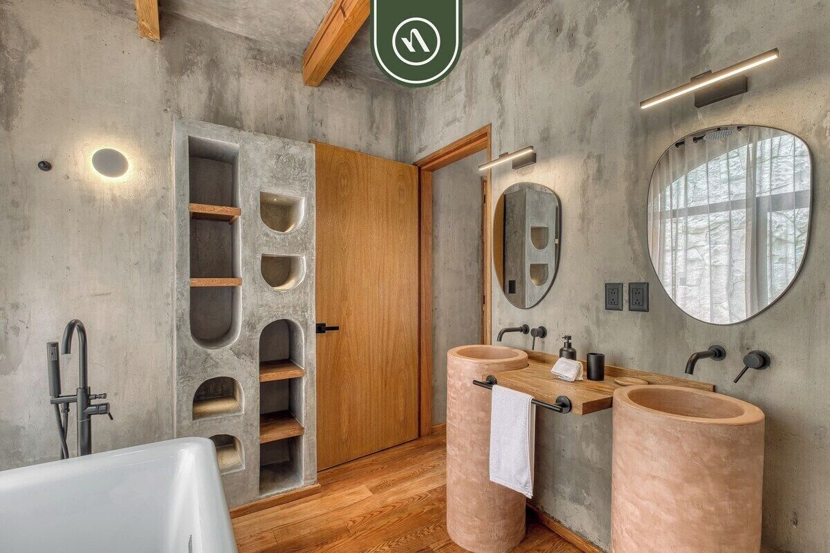 A bathroom with a concrete wall, wooden door, two sinks, a mirror, and a bathtub.