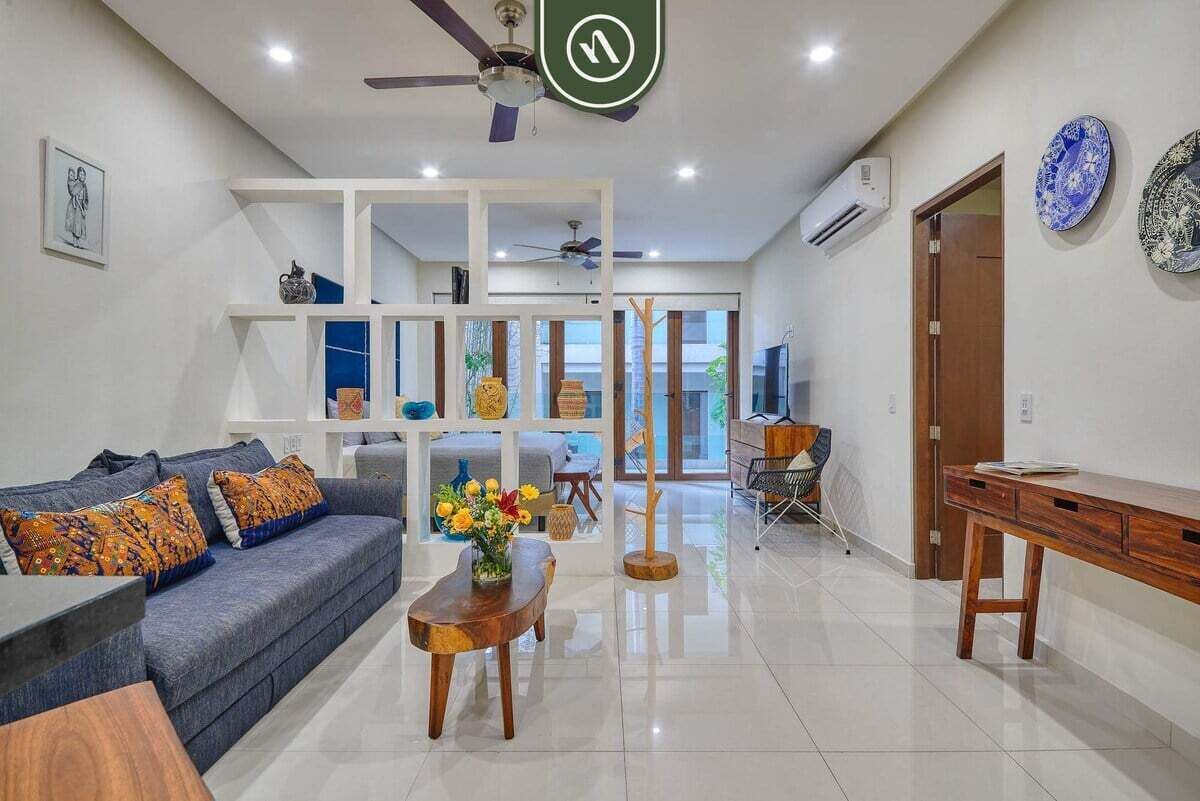 Ein modernes Wohnzimmer mit einer Couch, Couchtisch und Dekoration. Es gibt einen Essbereich mit Tisch und Stühlen, sowie einen Holztisch. Der Raum verfügt über einen Deckenventilator und eine Klimaanlage.