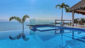 An infinity pool with a clear view of the ocean, a thatched-roof structure, and palm trees. - Luxury Oceanfront 3 BR Condo With Balcony (La Cruz de Huanacaxtle)
